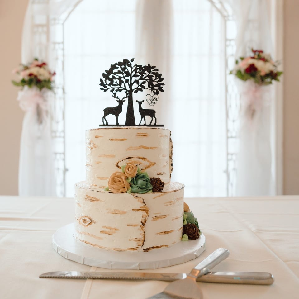 Two-tier birchwood-style wedding cake decorated with pastel flowers and a tree-and-deer topper, displayed beside serving utensils.