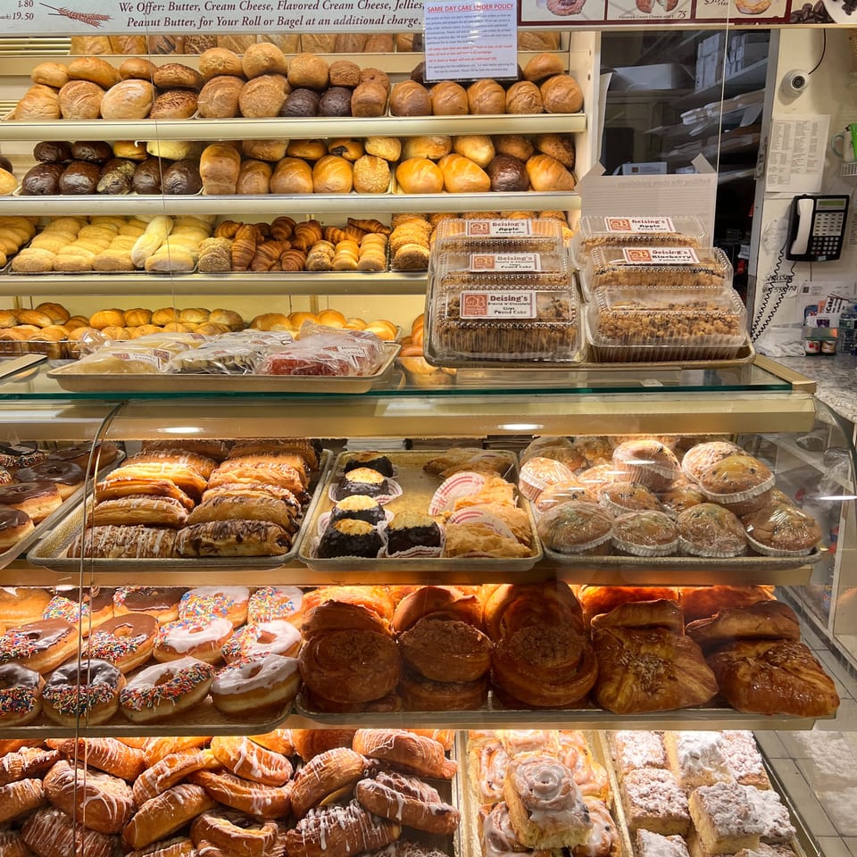 A glass bakery case displays rows of breads, bagels, pastries, donuts, cakes, and packaged pound cakes beneath menu boards.