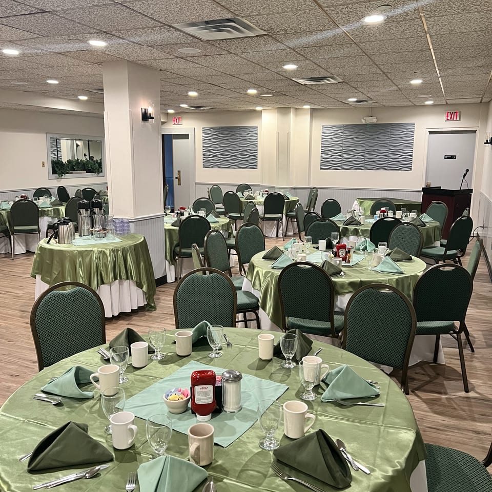 Spacious restaurant dining room with green tablecloths, neatly arranged place settings, dark green chairs, and a small podium.