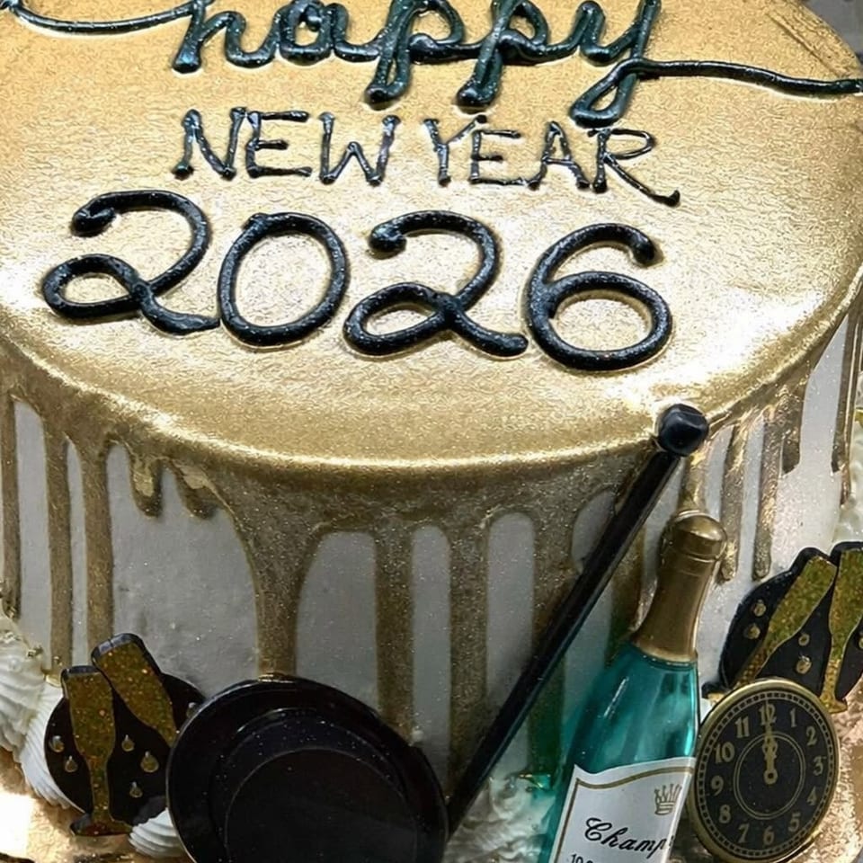 Gold-and-white celebration cake decorated with black lettering reading “Happy New Year 2026,” champagne bottle, clock, and party accents.