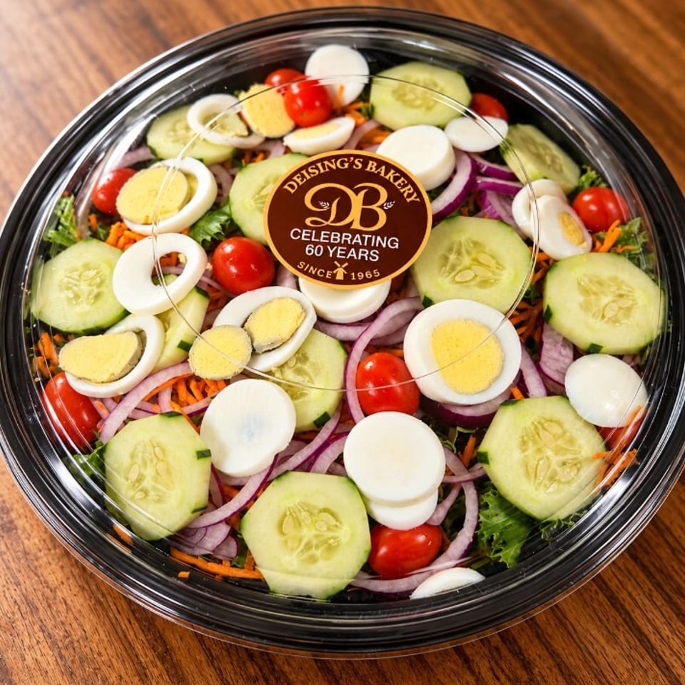 Overhead view of a garden salad with cucumbers, cherry tomatoes, carrots, red onions, lettuce, and sliced eggs in a clear container.