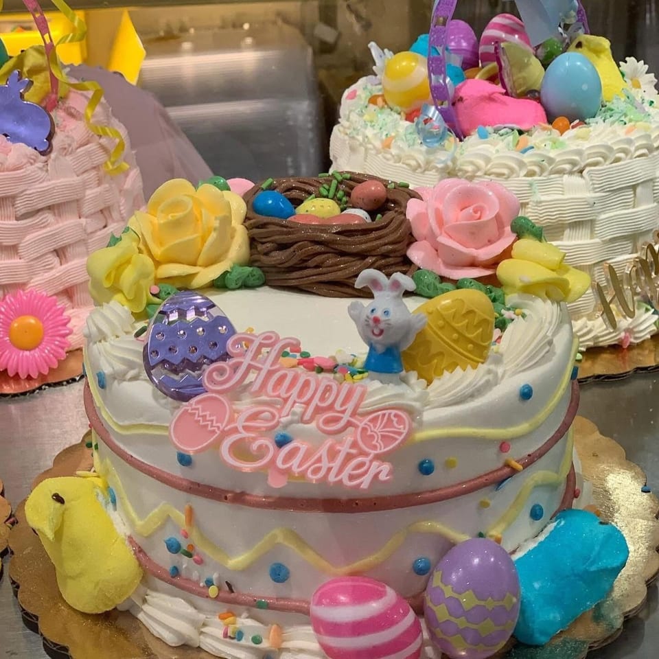 Colorful Easter cake decorated with pastel eggs, flowers, a bunny, chocolate nest, and pink “Happy Easter” icing.