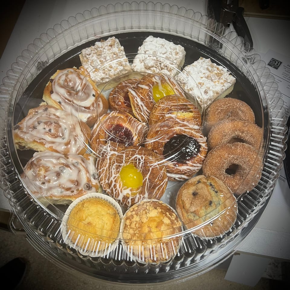 Assorted glazed pastries, fruit-filled danishes, cinnamon rolls, powdered bars, muffins, and sugar-coated donuts arranged in a plastic tray.