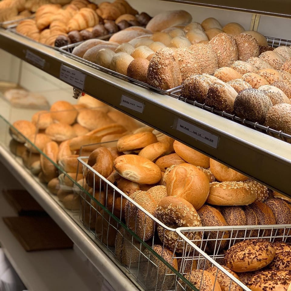 Shelves display assorted freshly baked breads and bagels, including sesame, poppy seed, and oat-topped varieties.