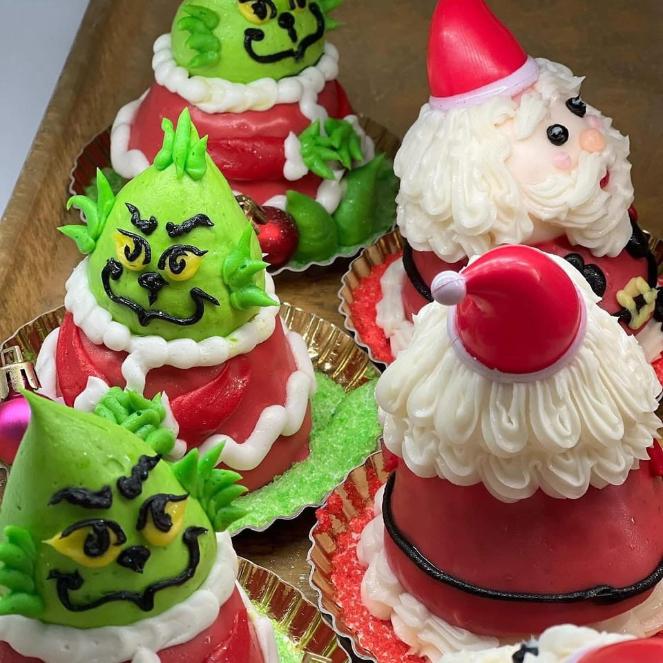 Colorful Christmas cupcakes decorated as Grinch characters and Santa Claus, with red-and-white frosting and intricate facial details.