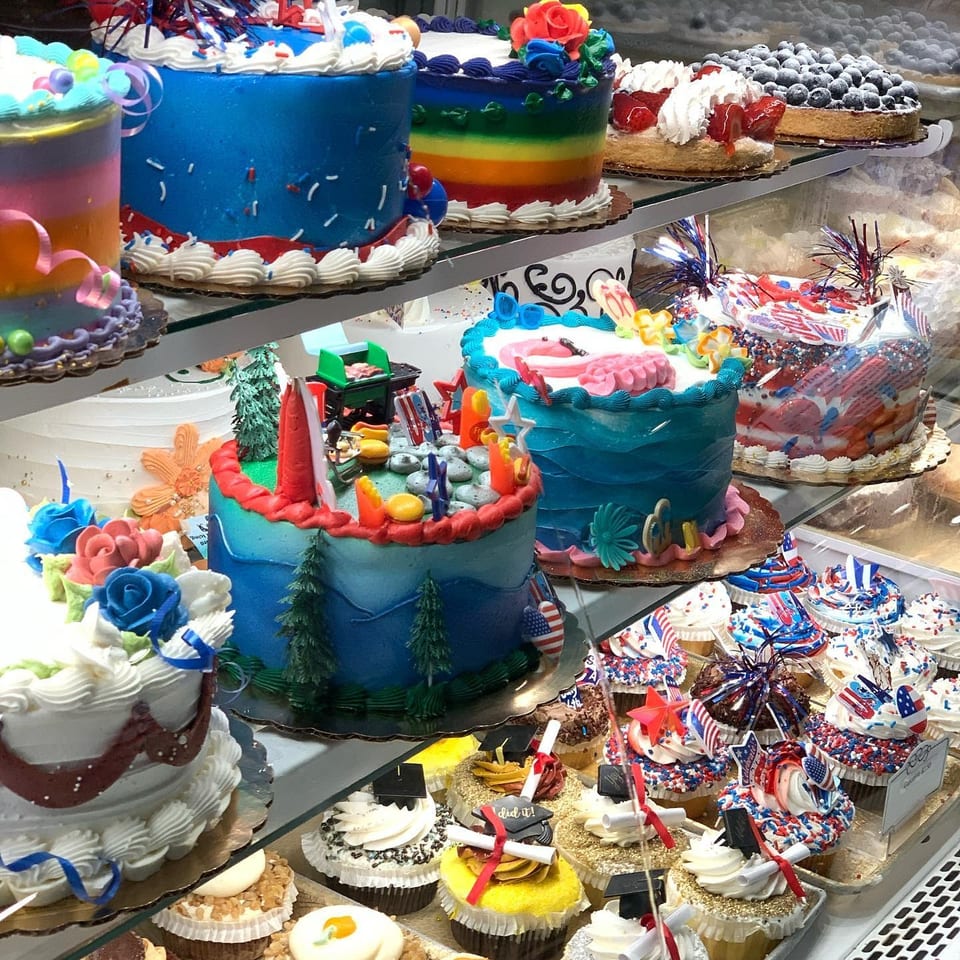 Colorful decorated cakes and cupcakes fill glass bakery display shelves, including rainbow layers, berries, flowers, and patriotic sprinkles.